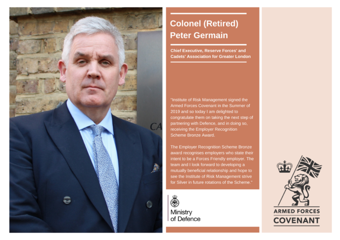 Colonel (Retired) Peter Germain, a distinguished Army veteran in a suit, stands in front of a brick wall next to a display highlighting his achievements with the Armed Forces Covenant and Ministry of Defence.