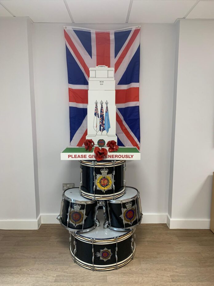 A display of British Army and Royal Navy drums stacked below a donation box and poppies, with a large Union Jack flag hung on the wall behind them.