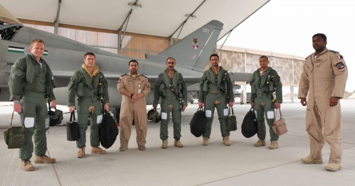 Six airmen in flight suits and one Army veteran in a tan uniform stand in front of a military jet under a canopy, posing for a group photo on a concrete surface.