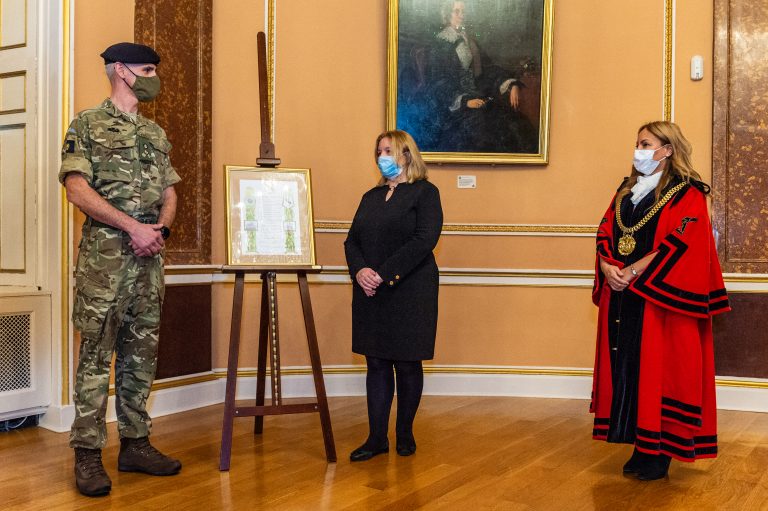 A Royal Navy soldier, a woman in black, and a woman in a red ceremonial robe stand beside a framed picture on an easel in an ornate room. All are wearing face masks.