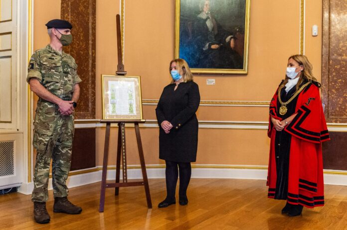 A Royal Navy soldier, a woman in black, and a woman in a red ceremonial robe stand beside a framed picture on an easel in an ornate room. All are wearing face masks.