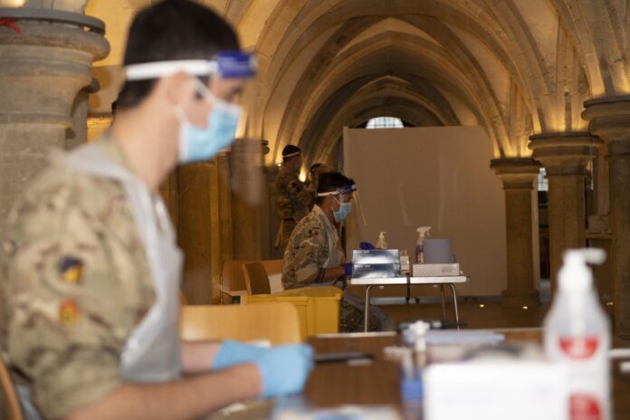 Veteran soldiers wearing masks and face shields sit at tables with medical supplies in a large stone hall, preparing for a military medical or testing procedure.