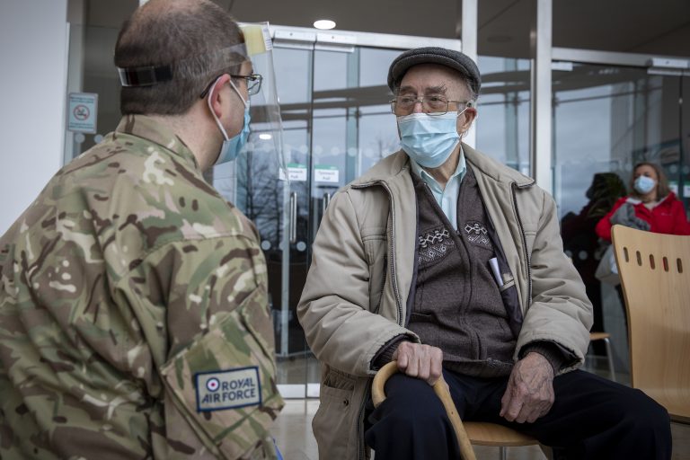 An elderly man wearing a mask sits with a cane, speaking to a uniformed Royal Air Force member indoors. Another masked person, possibly from the Army or Royal Navy, stands in the background.