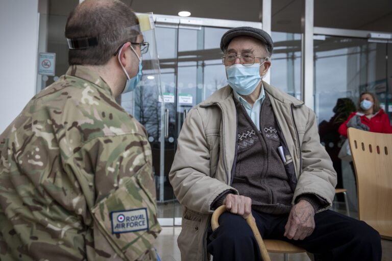 An elderly man wearing a mask sits with a cane, speaking to a uniformed Royal Air Force member indoors. Another masked person, possibly from the Army or Royal Navy, stands in the background.