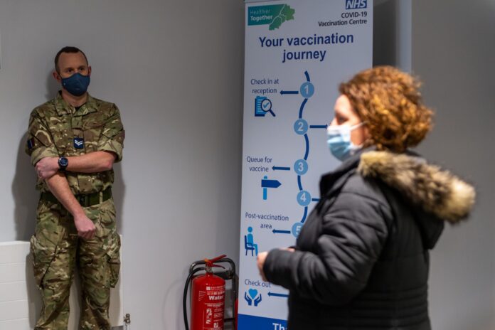 A veteran in military uniform stands by a wall as a woman wearing a mask approaches a COVID-19 vaccination centre sign displaying the vaccination process steps.