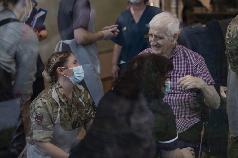 A healthcare worker in uniform and mask kneels beside an elderly man in a wheelchair, both surrounded by other masked staff in a medical resettlement setting.