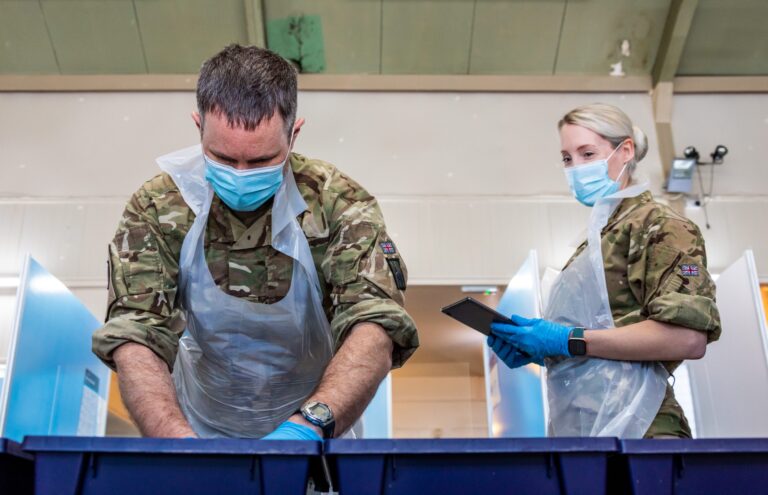 Two uniformed individuals, possibly military or Royal Navy personnel, wearing face masks and gloves, sort through documents at a table indoors, with partitioned booths in the background.