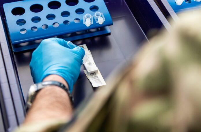 A person wearing a blue glove prepares a medical test strip and lab vials on a tray, with blue test tube racks in the background—part of a resettlement program for former Royal Navy personnel transitioning to civilian healthcare roles.