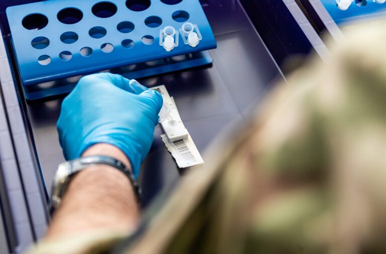 A person wearing a blue glove prepares a medical test strip and lab vials on a tray, with blue test tube racks in the background—part of a resettlement program for former Royal Navy personnel transitioning to civilian healthcare roles.