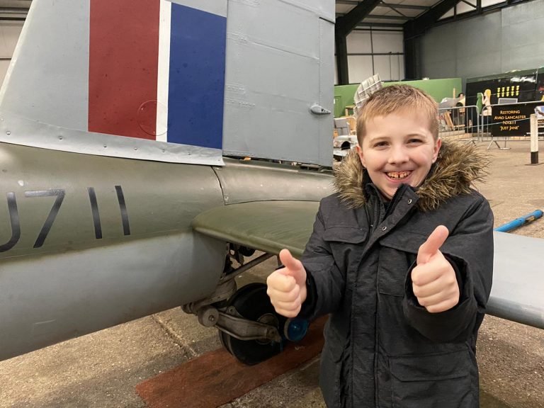 A young boy in a black coat stands beside an aircraft tail, smiling and giving two thumbs up inside a hangar, proudly posing like a Royal Navy veteran.