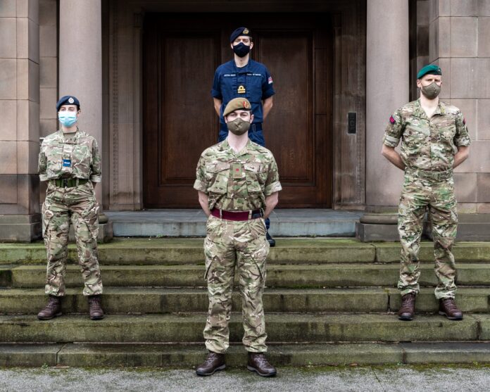 Four uniformed Army personnel wearing face masks stand spaced apart on steps in front of a large wooden door and stone building.