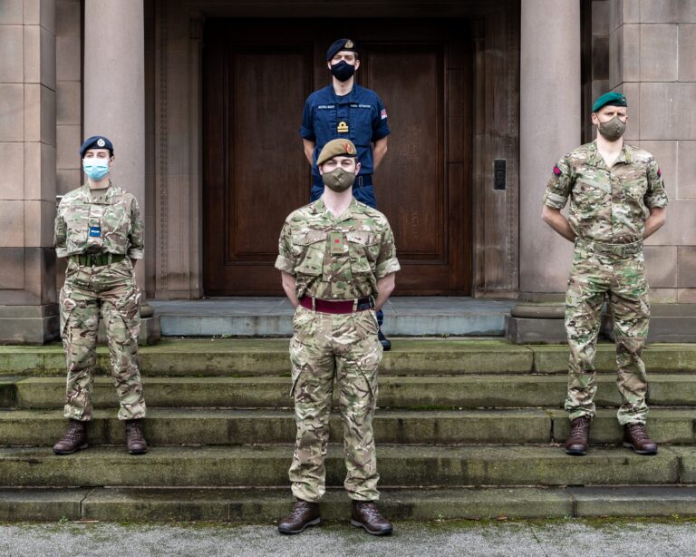 Four uniformed Army personnel wearing face masks stand spaced apart on steps in front of a large wooden door and stone building.