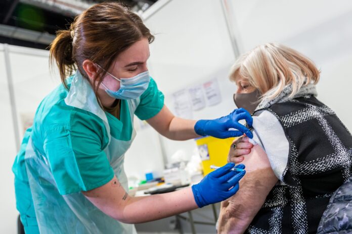A healthcare worker in scrubs and gloves administers a vaccine injection to an older woman’s upper arm at a medical facility, supporting her resettlement into safer, healthier living.