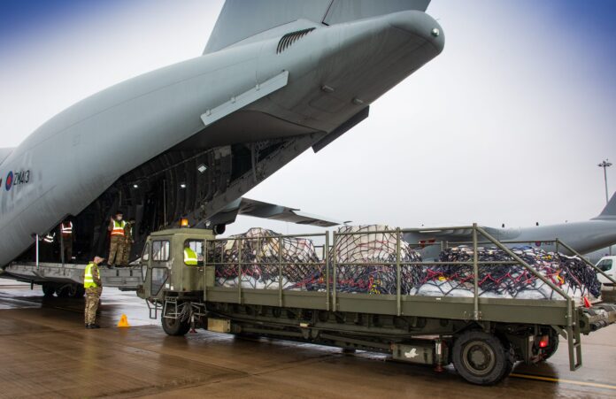Cargo is being loaded onto a military transport aircraft from a truck on an airport tarmac, with veteran personnel supervising the operation.