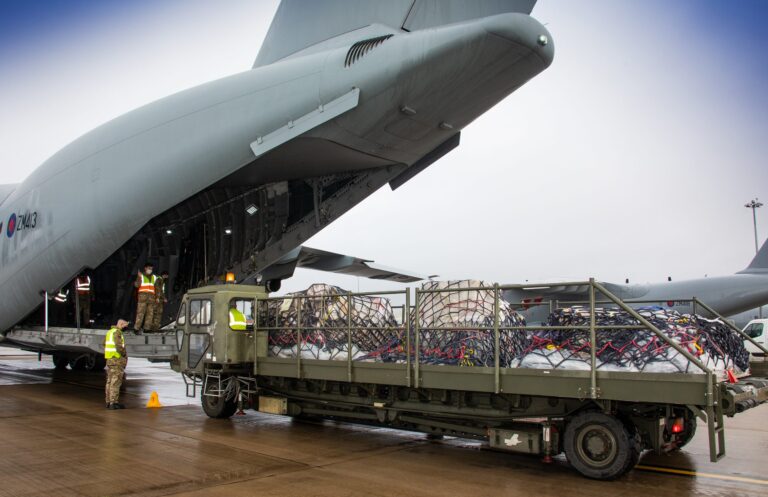 Cargo is being loaded onto a military transport aircraft from a truck on an airport tarmac, with veteran personnel supervising the operation.