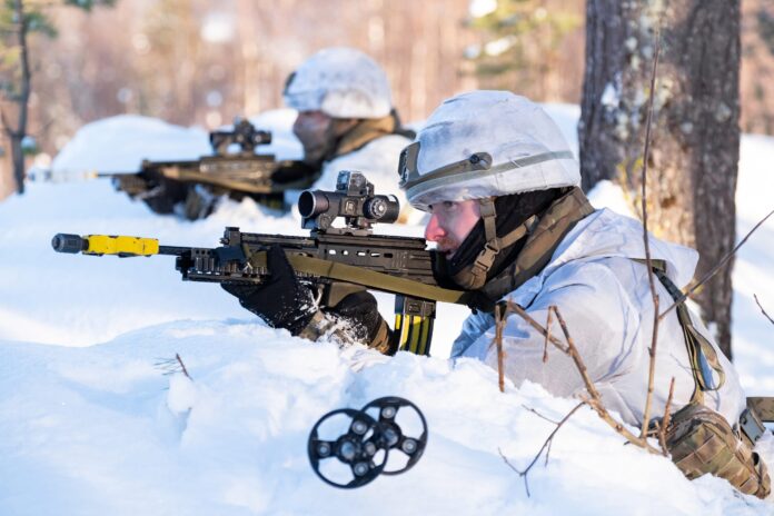 Two Army soldiers in white winter camouflage aim rifles while positioned in snowy terrain, with trees and snow in the background.