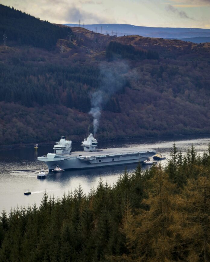 A large Royal Navy aircraft carrier with escort boats sails through a narrow body of water surrounded by forested hills, with smoke rising from the ship.
