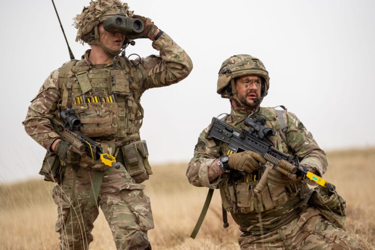 Two Army soldiers in camouflage uniforms and helmets, one using binoculars and the other holding a rifle, kneel in a grassy field during a military exercise.