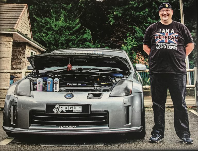 A man stands next to a silver car with its hood open, displaying the engine and several bottles; he is smiling and wearing a military veteran-themed shirt and cap.