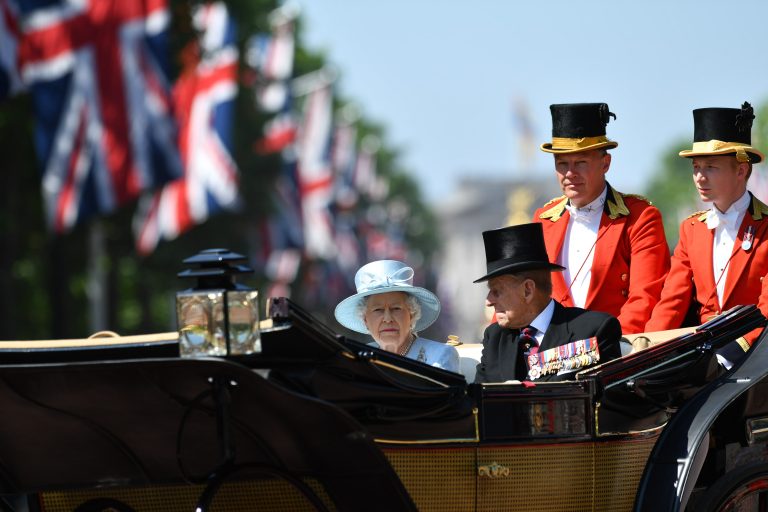 A woman in a light blue hat and a man in uniform, possibly a veteran, ride in a horse-drawn carriage, accompanied by three men in red coats and top hats, with Union Jack flags lining the street.