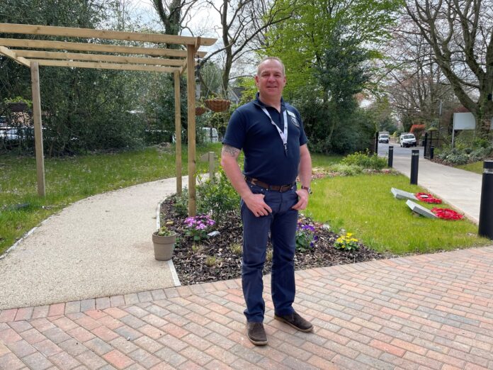 A man in a navy polo shirt and jeans stands on a brick walkway beside a flowerbed and grassy area, with trees and parked cars in the background, reminiscent of a Royal Navy pathfinder enjoying some time off duty.