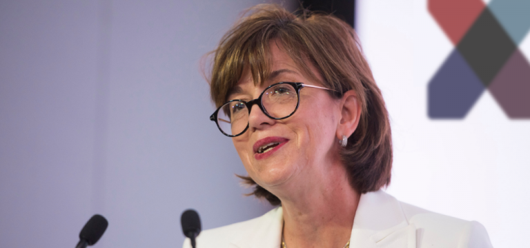 A woman with short brown hair and glasses, possibly a veteran, speaks at a podium with two microphones, wearing a white blazer; a blurred geometric design is in the background.