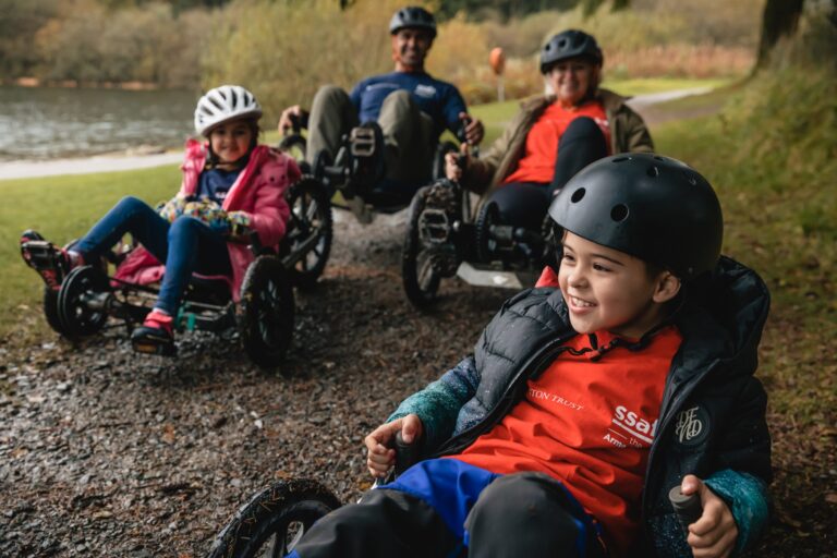 Four people wearing helmets ride Pathfinder recumbent trikes on a gravel path beside a lake, surrounded by trees and grass.