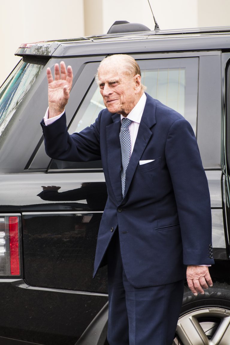 A Royal Navy veteran in a navy suit waves while standing next to a black SUV.