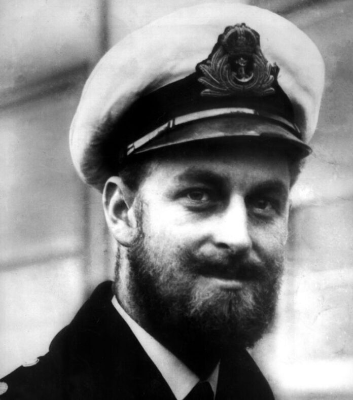 A man in a Royal Navy officer's uniform and cap with an emblem, sporting a beard, is looking at the camera and smiling slightly.