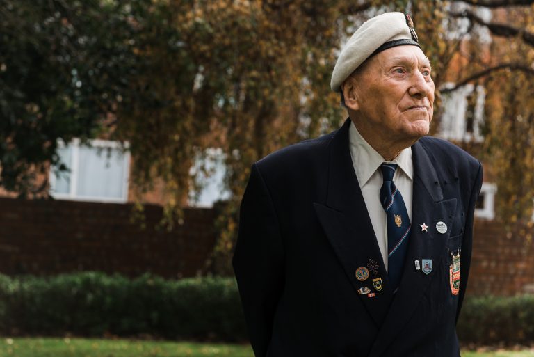 An elderly military veteran in a suit and beret with badges stands outdoors, looking to the side, with trees and a brick wall in the background.