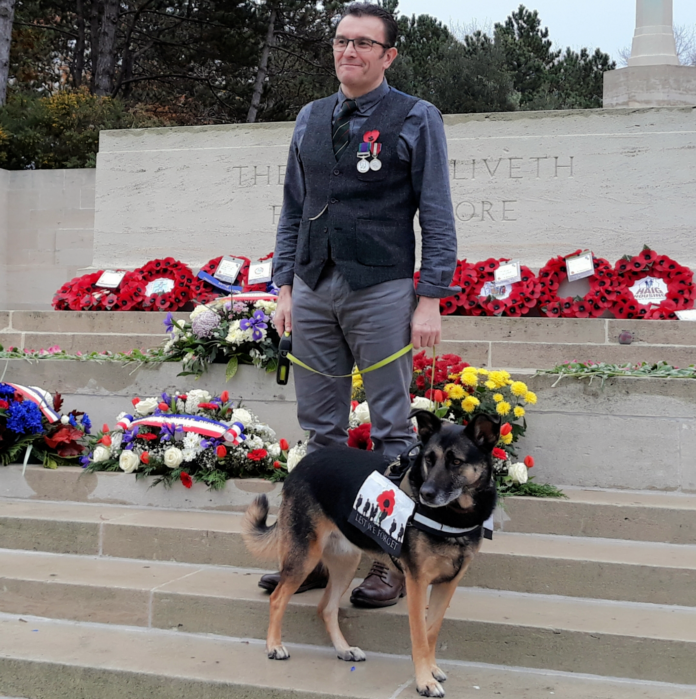 A man wearing medals stands on steps beside a German Shepherd dog in a poppy vest; wreaths of flowers are displayed on the steps and a military memorial wall is behind them.