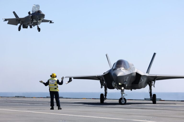 A crew member, possibly a veteran, signals to a parked fighter jet on an aircraft carrier as another jet approaches for landing in the background.