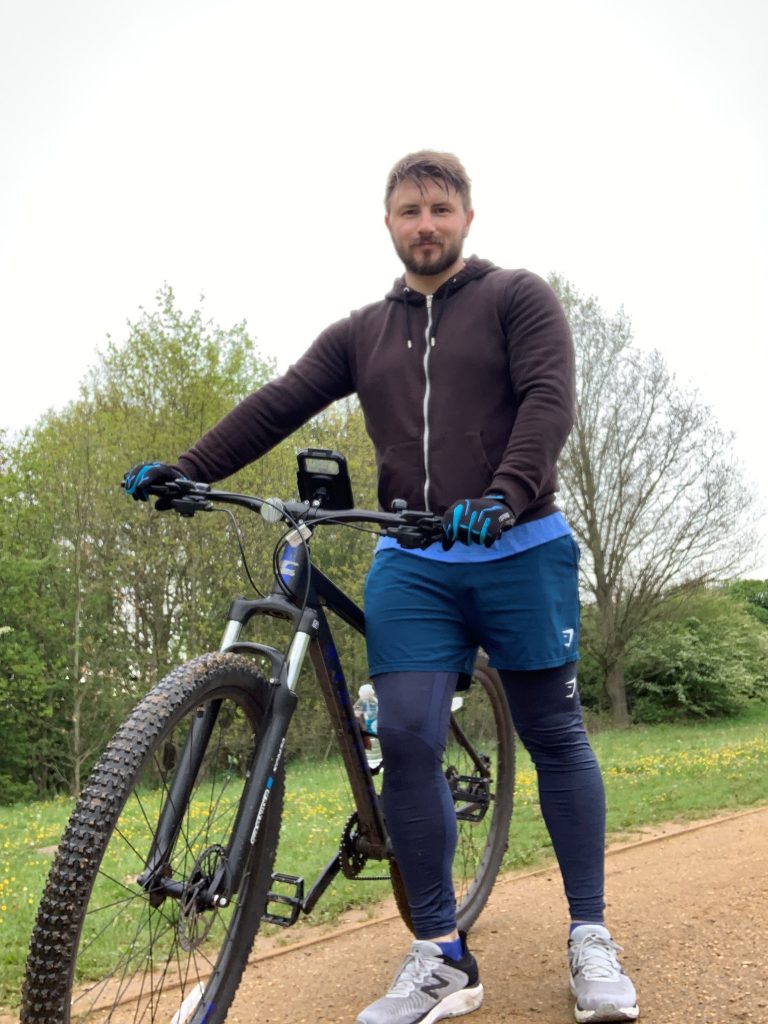 A man stands outdoors on a dirt path holding a mountain bike, wearing a hoodie, shorts, leggings, and gloves—like a true pathfinder—with trees and grass in the background.
