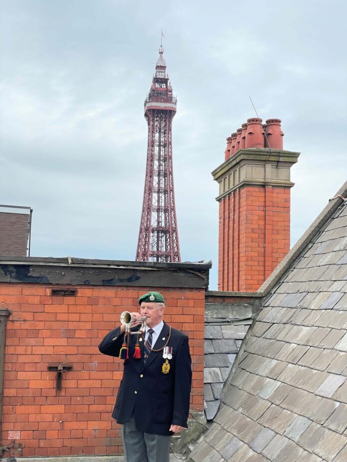 A military man in uniform with medals plays a bugle on a rooftop, with Blackpool Tower and a brick chimney visible in the background under a cloudy sky.