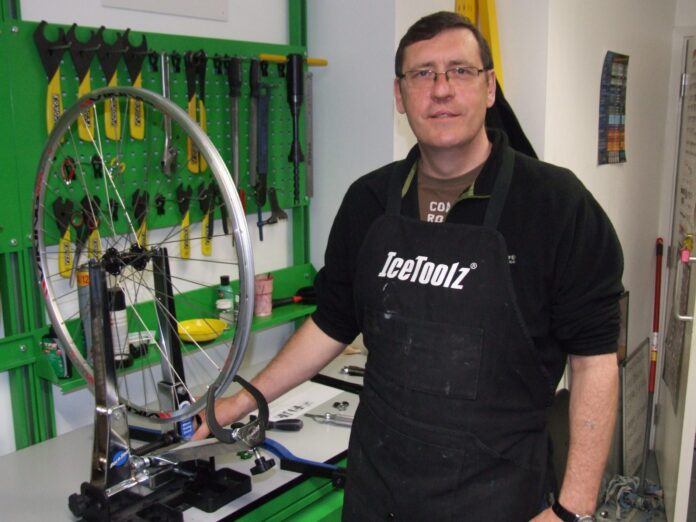 A veteran wearing glasses and a black apron stands beside a bicycle wheel truing stand in a workshop, tools neatly arranged on the wall behind him.