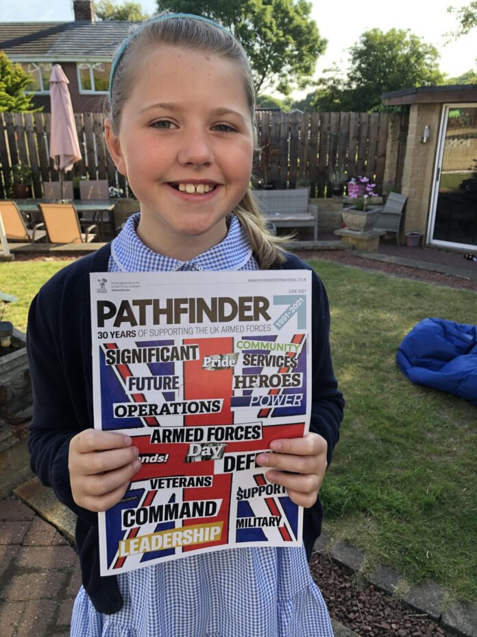 A smiling girl in a blue school uniform holds up a copy of Pathfinder magazine outdoors in a garden setting, proudly showcasing her interest in the stories of Royal Navy veterans.