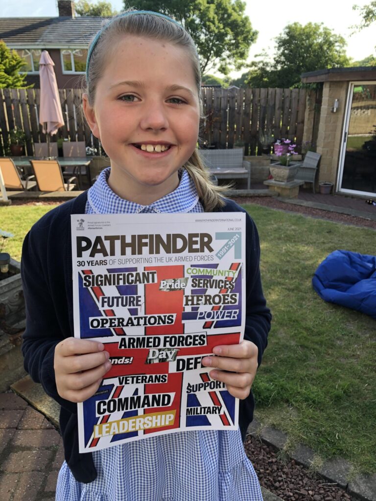 A smiling girl in a blue school uniform holds up a copy of Pathfinder magazine outdoors in a garden setting, proudly showcasing her interest in the stories of Royal Navy veterans.