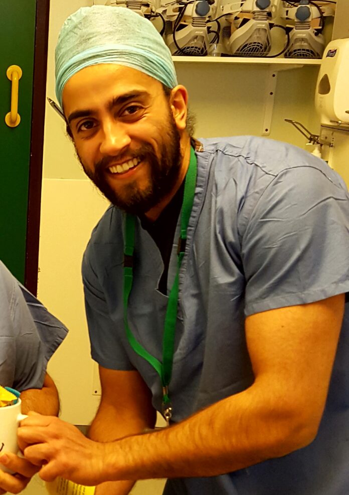 A military veteran in blue medical scrubs and a surgical cap smiles at the camera in a hospital or clinical setting.