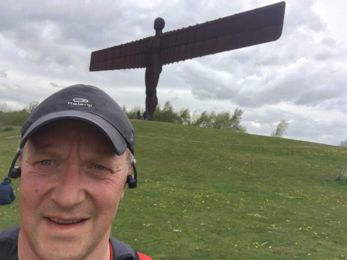 A veteran takes a selfie outdoors with the large Angel of the North sculpture in the background under a cloudy sky.