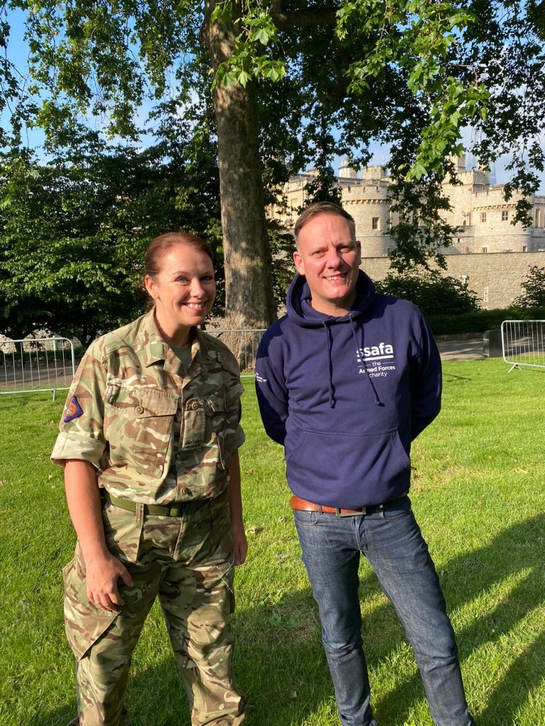 A woman in Royal Navy uniform stands next to a man in a blue hoodie on a grassy area, with trees and a stone building in the background, symbolizing the spirit of resettlement.