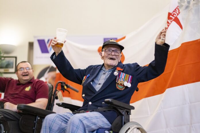 An elderly military man in a wheelchair raises a drink and waves in front of an England flag, wearing medals and a navy blazer; another man sits nearby, smiling.