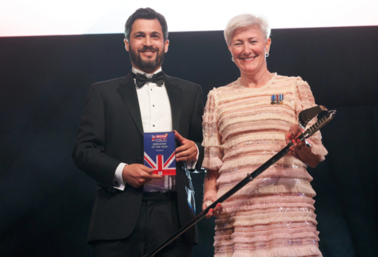 A man in a tuxedo holds an award plaque while standing next to a woman in a formal dress holding a ceremonial sword, both smiling at a Royal Navy event.