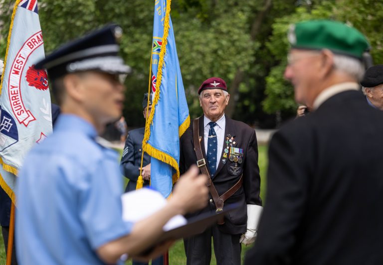 An elderly man wearing military medals and a beret stands outdoors with others, near flags, during a commemorative military event.