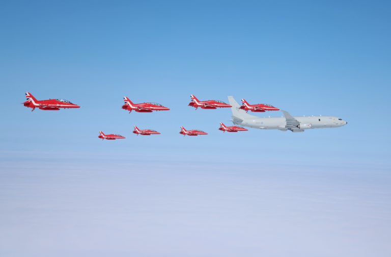 Nine red jets and one larger white military aircraft fly in formation above a blanket of clouds under a clear blue sky.