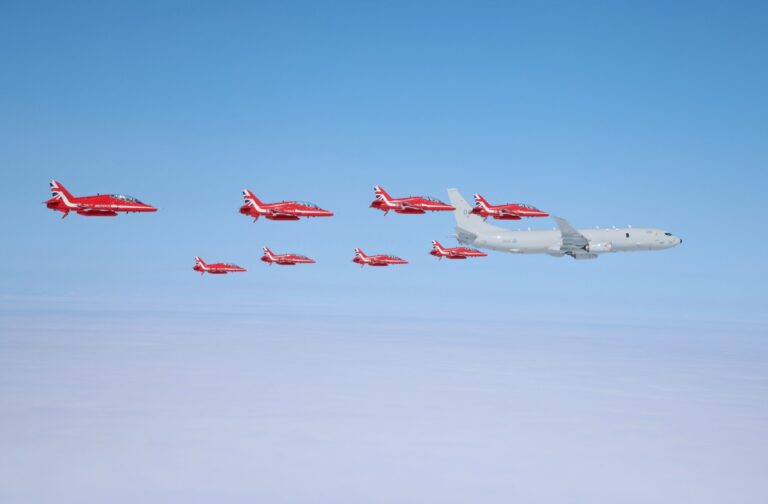 Nine red jets and one larger white military aircraft fly in formation above a blanket of clouds under a clear blue sky.