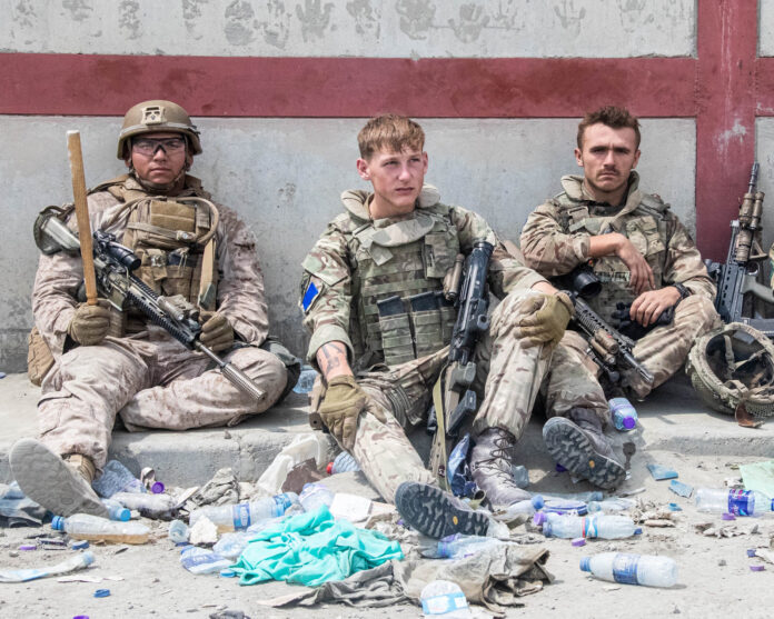 Three armed military soldiers in camouflage sit on a curb against a wall, surrounded by discarded water bottles and debris, appearing to wait during a resettlement operation.