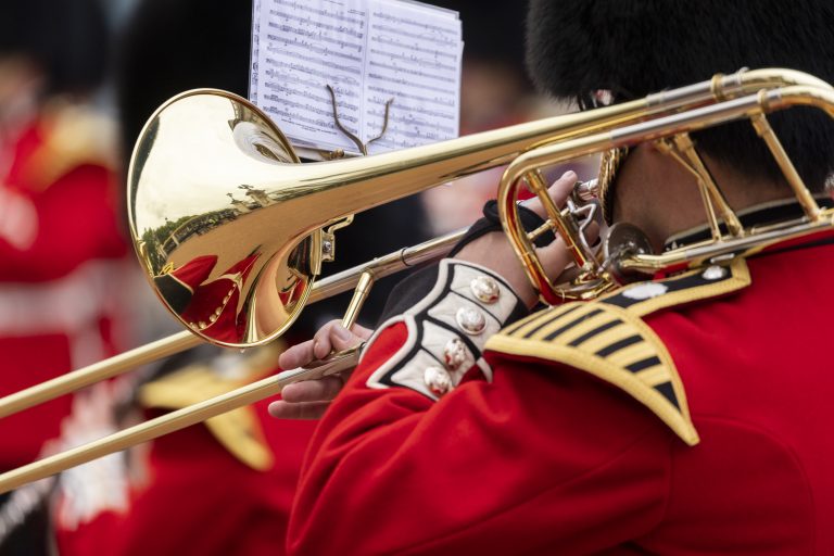 A musician in a red Army uniform plays a trombone with sheet music attached, while other uniformed people are visible in the blurred background.