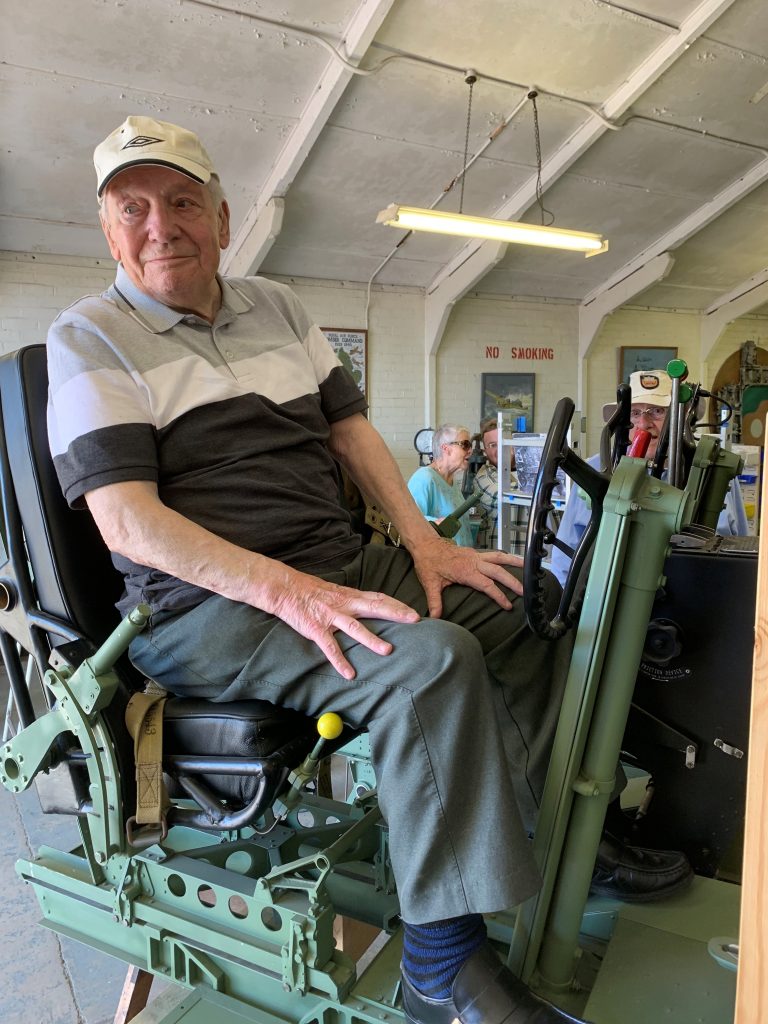 An elderly man in a striped shirt and cap sits on a mechanical chair indoors, exuding the quiet dignity of a Royal Navy veteran, while two other people work in the background.