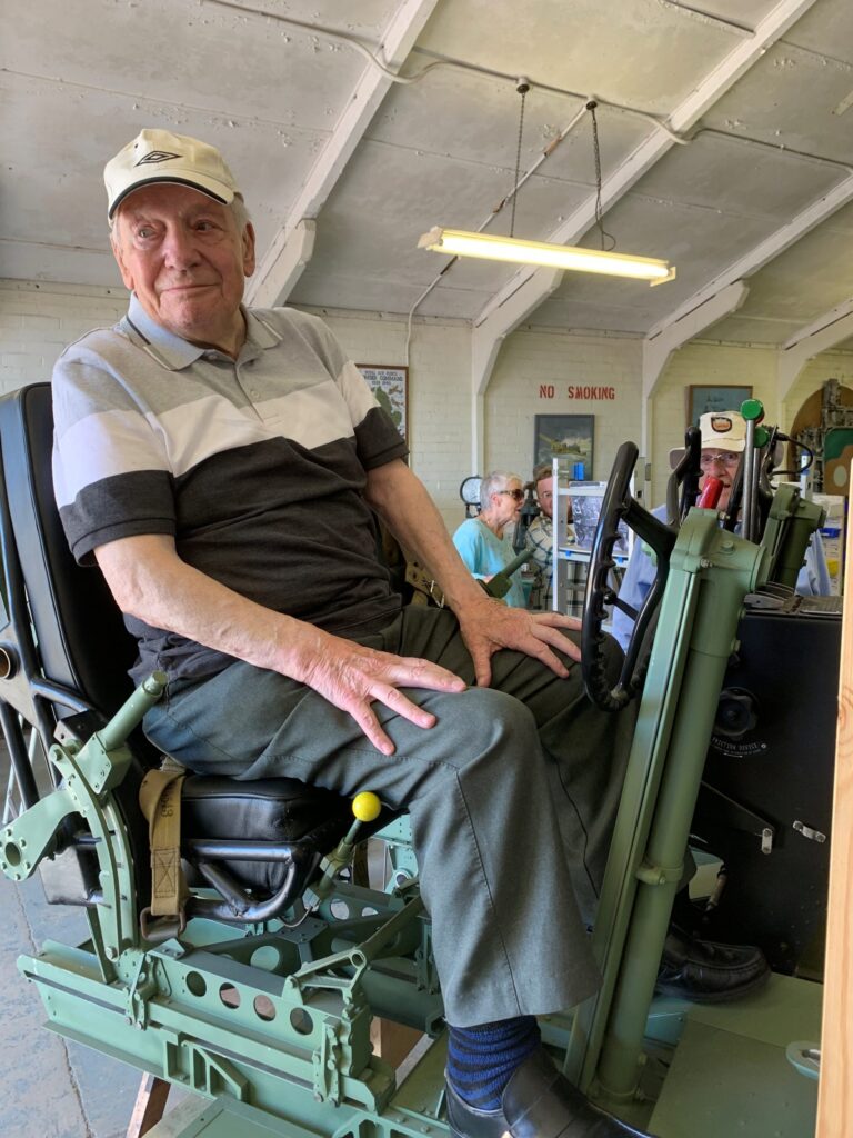 An elderly man in a striped shirt and cap sits on a mechanical chair indoors, exuding the quiet dignity of a Royal Navy veteran, while two other people work in the background.