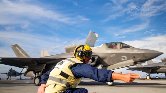 A veteran flight deck crew member directs a fighter jet during takeoff on an aircraft carrier under a partly cloudy sky.
